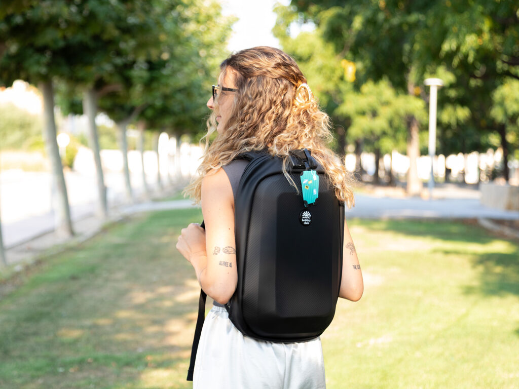 Chica de espaldas con mochila negra rígida de ALE-HOP equipada con un tag para maletas azul.