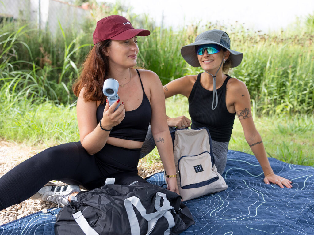 Mujeres con gorra deportiva y sombrero pescador descansando sobre una manta de picnic con mochila plegable ALE-HOP.