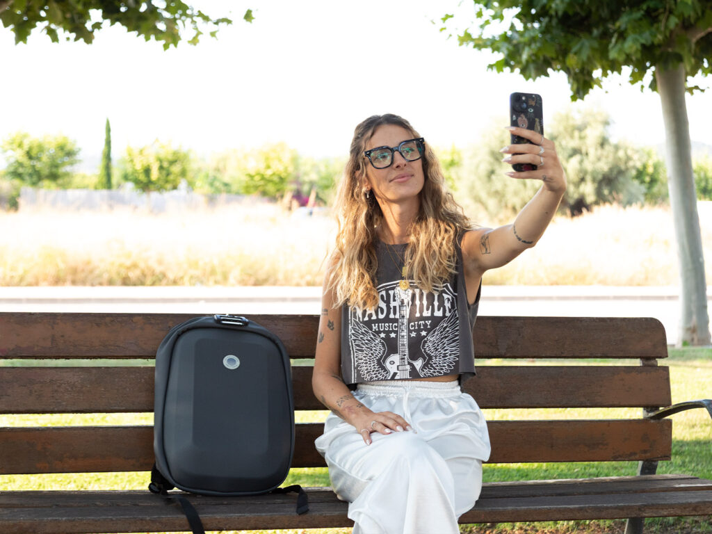 Chica haciéndose un selfie en un banco junto a una mochila de viaje negra y accesorios de viaje de ALE-HOP.