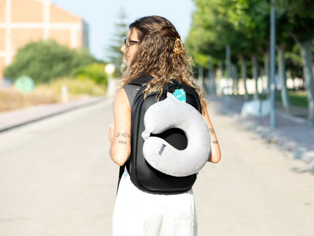 Chica de espaldas con mochila de viaje rígida de ALE-HOP y una almohada de viaje gris para el cuello.