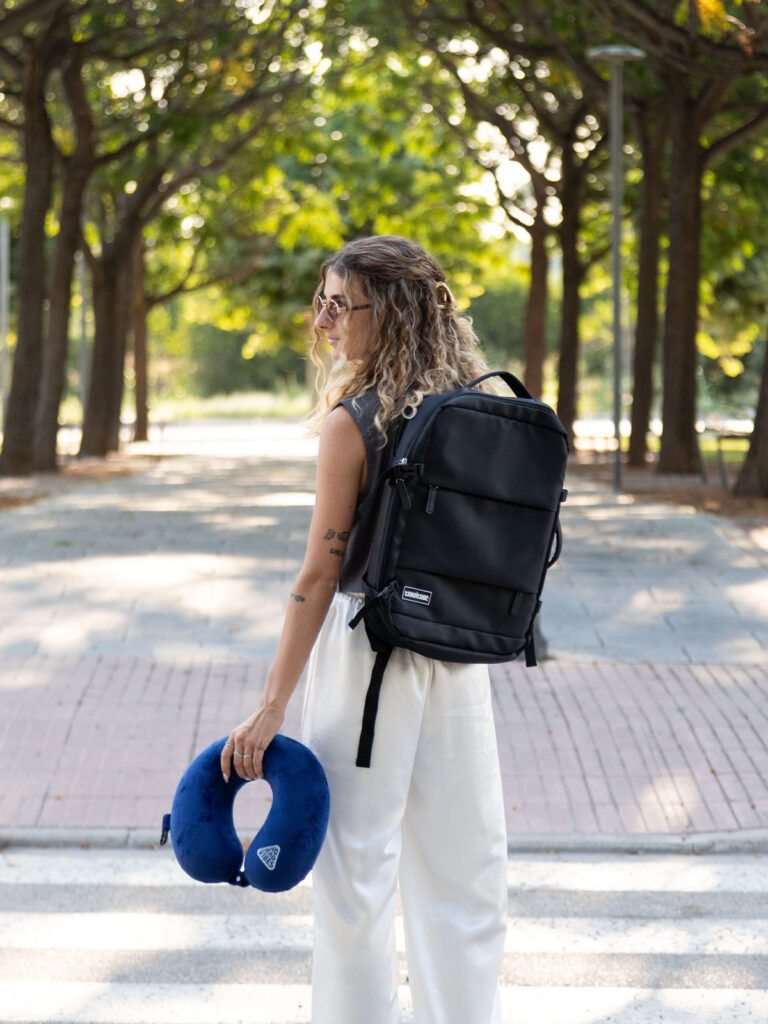 Chica de espaldas con mochila de equipaje de mano negra y almohada de cuello para viaje azul de ALE-HOP.