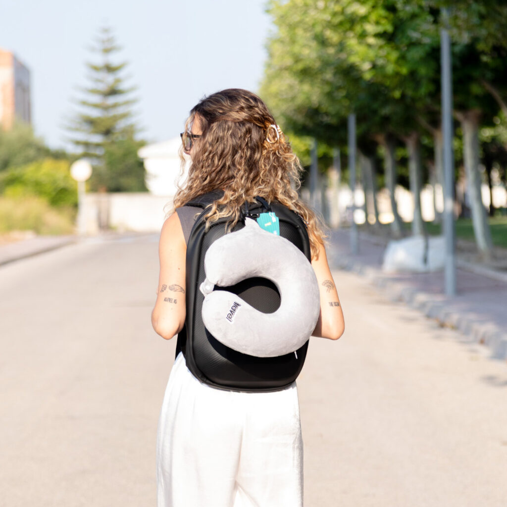 Chica de espaldas con mochila negra de ALE-HOP y una almohada de viaje gris para el cuello.