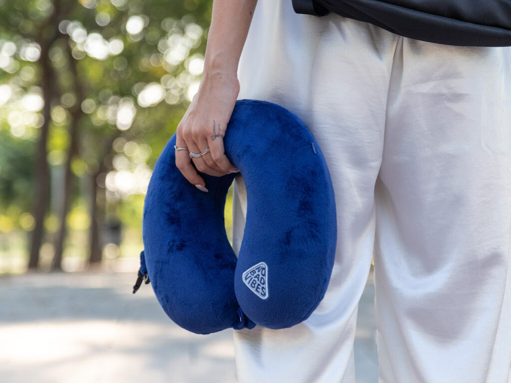 Detalle de mano sujetando una almohada cuello viaje azul de ALE-HOP con el parche No Bad Vibes.