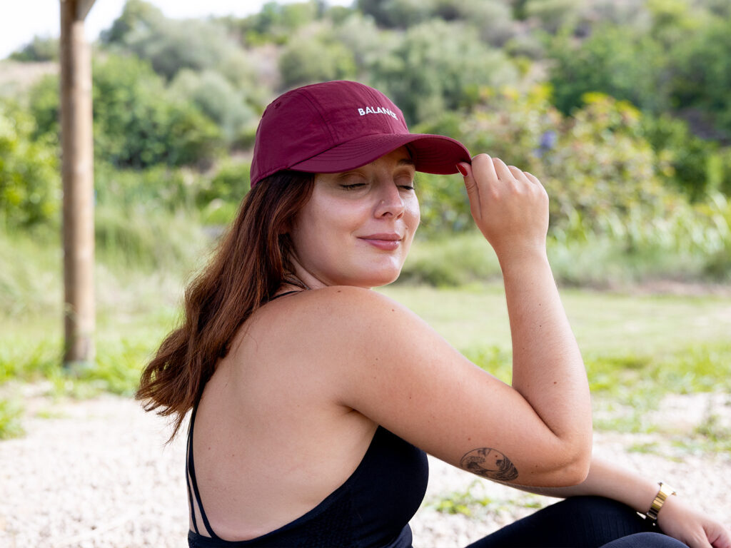 Mujer relajada al aire libre protegiéndose del sol con una gorra deportiva granate de la colección de verano de ALE-HOP.