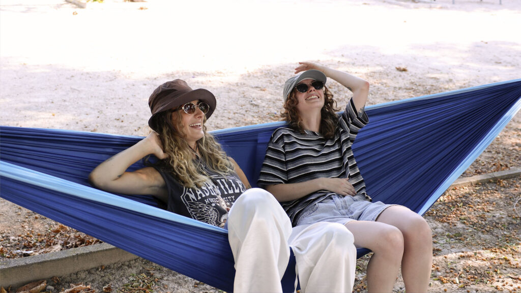 Dos amigas riendo en una hamaca colgante azul de ALE-HOP, con gorra y sombrero pescador.