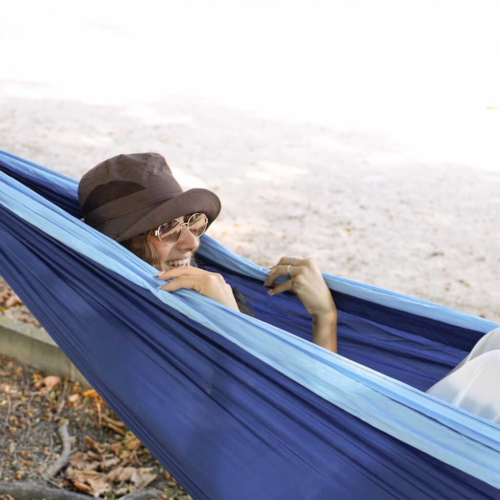 Chica relajándose en hamaca colgante de ALE-HOP con sombrero de playa y gafas de sol.