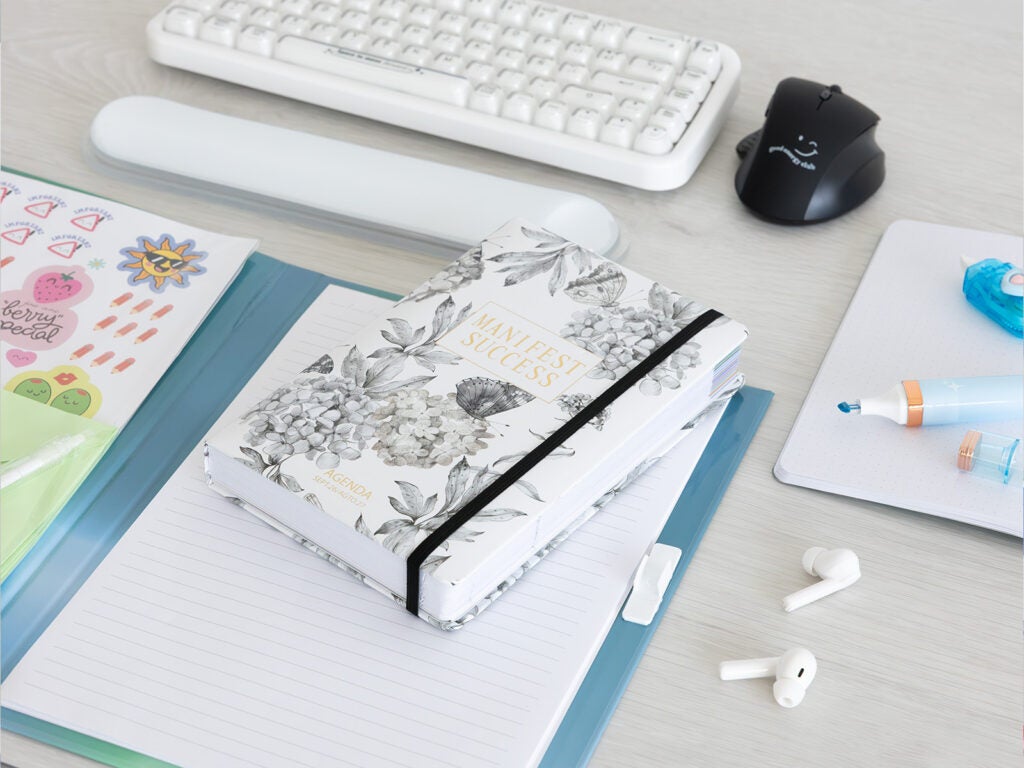 Agenda escolar 26/27 con diseño floral junto a teclado y ratón en mesa de trabajo.