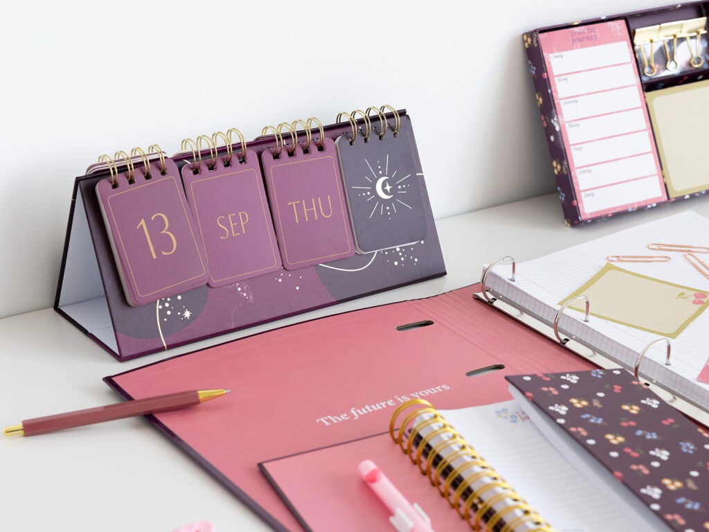 Calendario sobremesa perpetuo junto a carpeta de anillas y libreta de notas sobre escritorio.