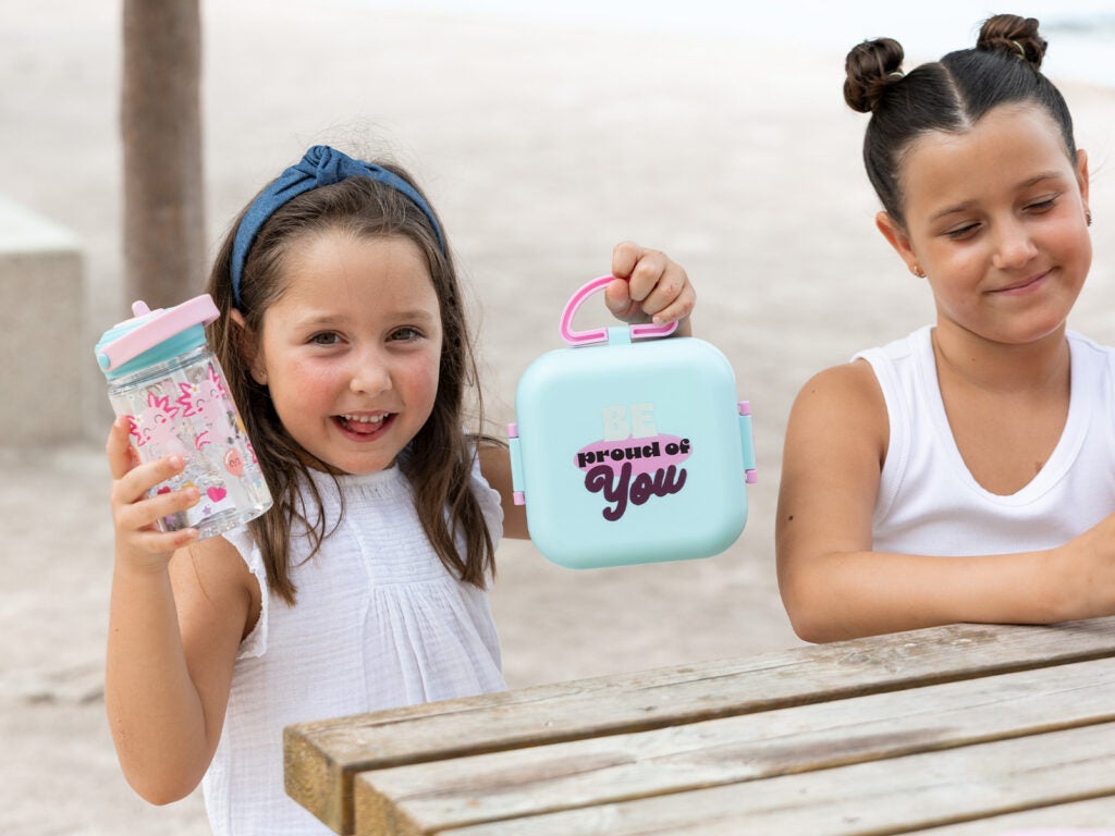 Niñas en la playa mostrando porta meriendas azul y botella de agua infantil para la vuelta al cole.
