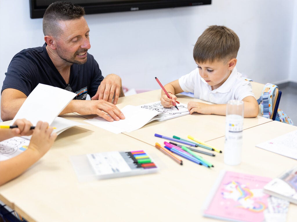 Profesor ayudando a alumno a colorear con rotuladores de colores en el aula escolar.